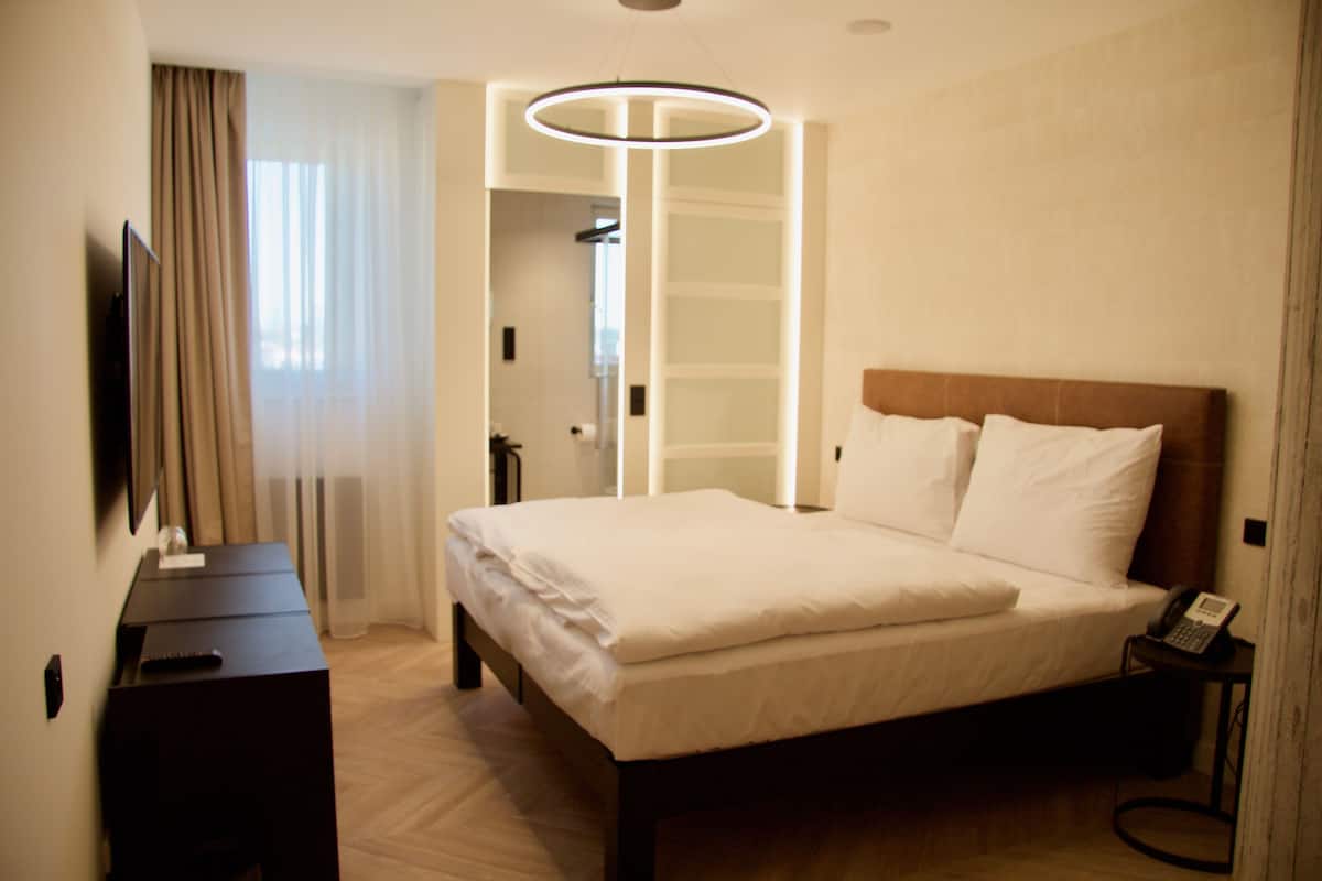 Habitación de hotel con cama, mesita de noche, lámpara y vistas al baño.