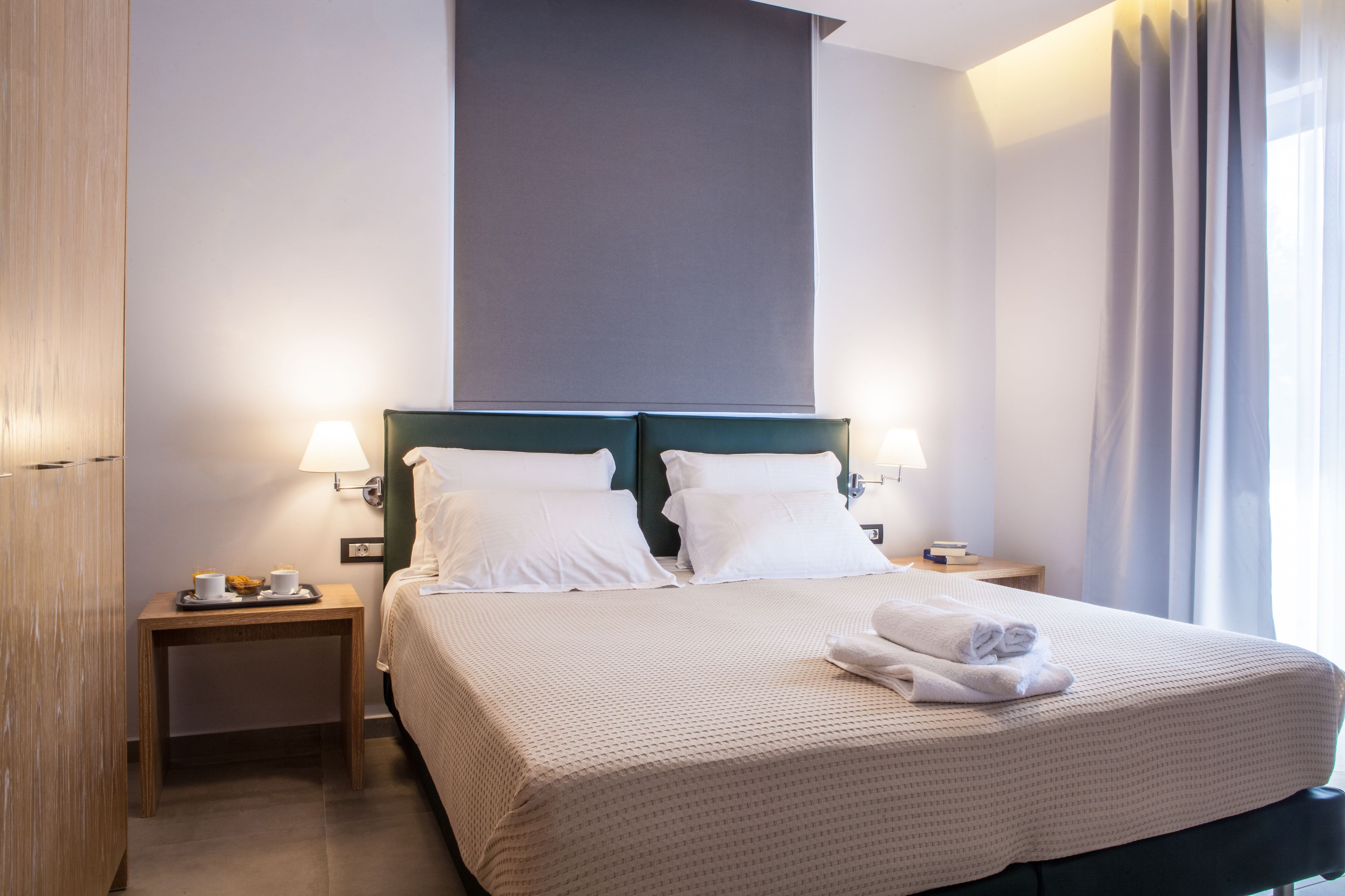 A neatly made bed with white linens, two bedside tables with lamps, and a large window with curtains.