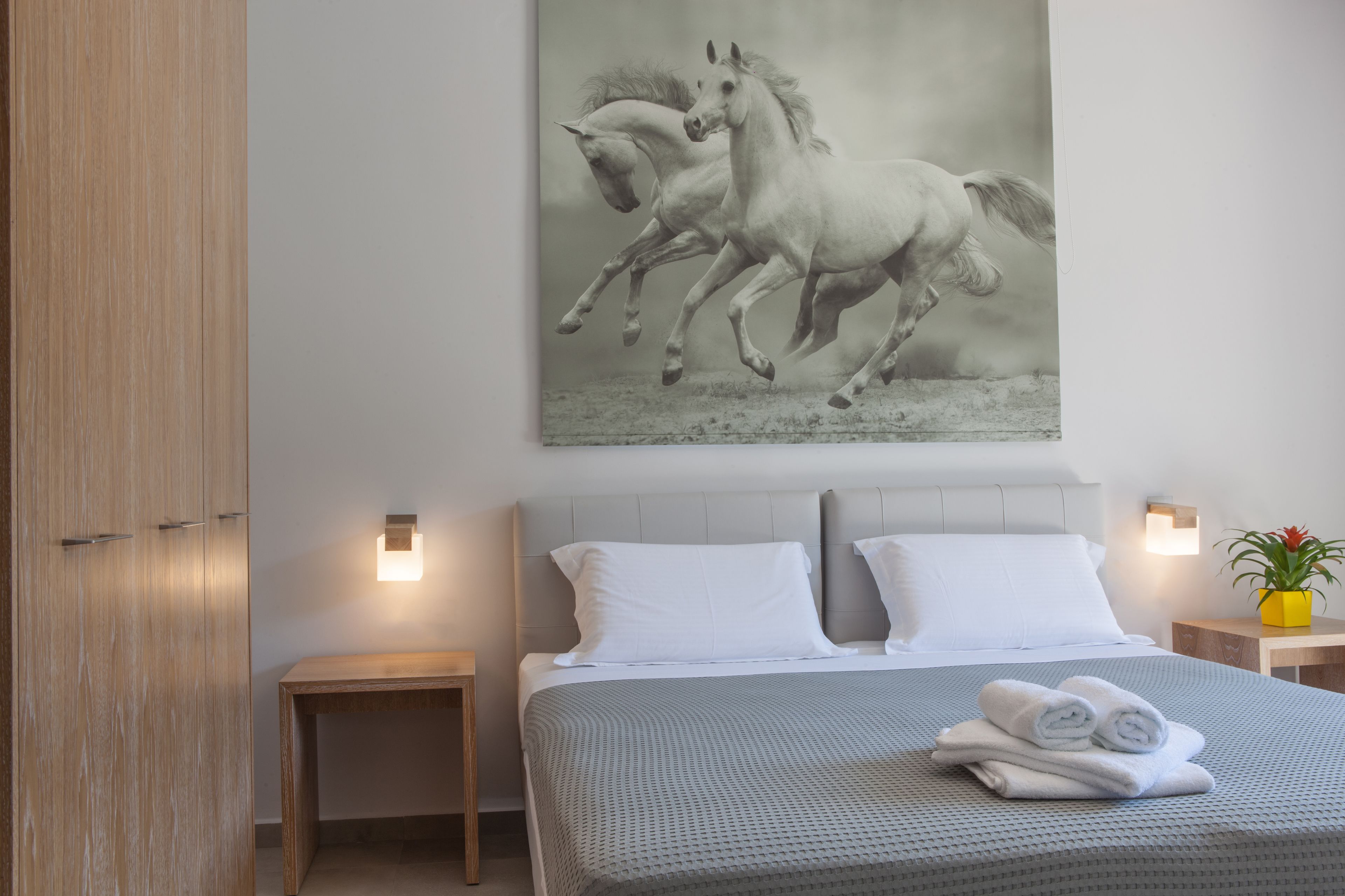 A bedroom with a large bed, two bedside tables, a nightstand, a wall-mounted light, and a painting of horses.