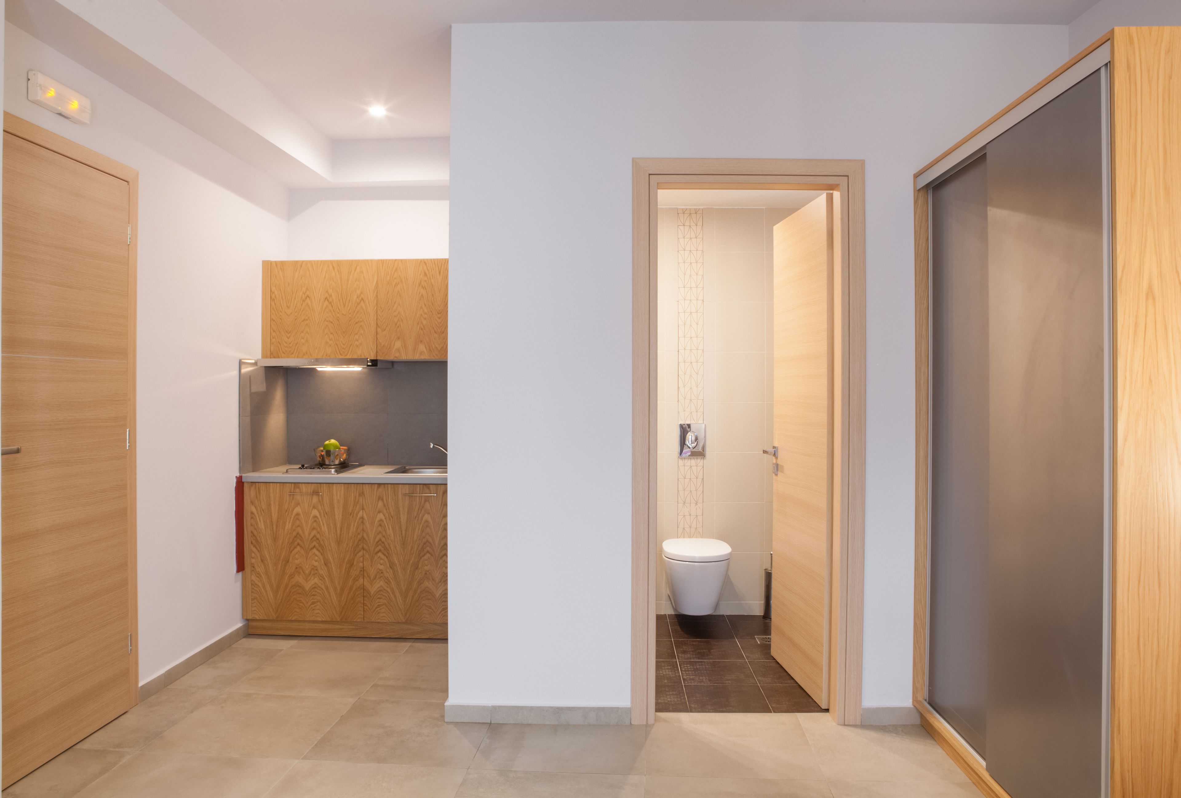 Una cocina moderna con armarios de madera y horno empotrado, una pequeña encimera con lavabo, y un inodoro en una habitación separada.