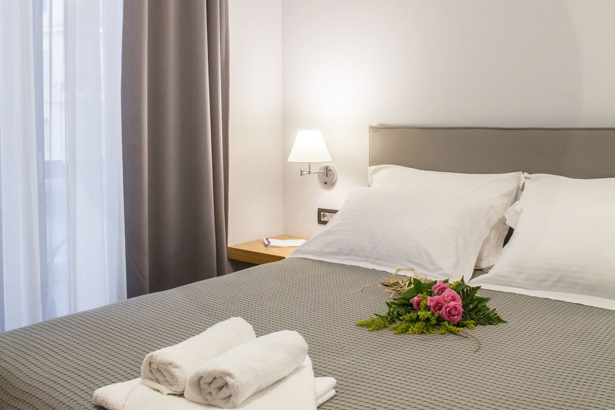 A neatly made bed with rolled towels and a floral arrangement, a bedside table with a lamp, and a window with sheer curtains.