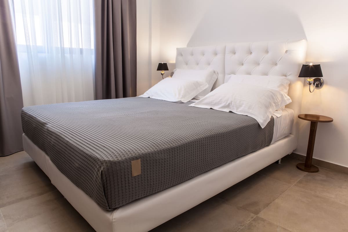 A neatly made bed with a grey patterned bedspread, white pillows, and a tufted headboard. There are two bedside tables with lamps, and a large window with sheer curtains.