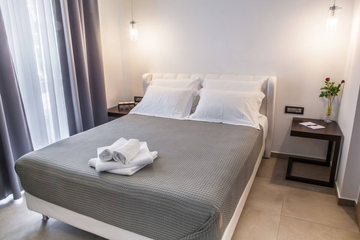 A neatly made bed with a grey and white bedspread, white pillows, and a folded white towel on the bed. There is a small bedside table with a vase of flowers and a book.