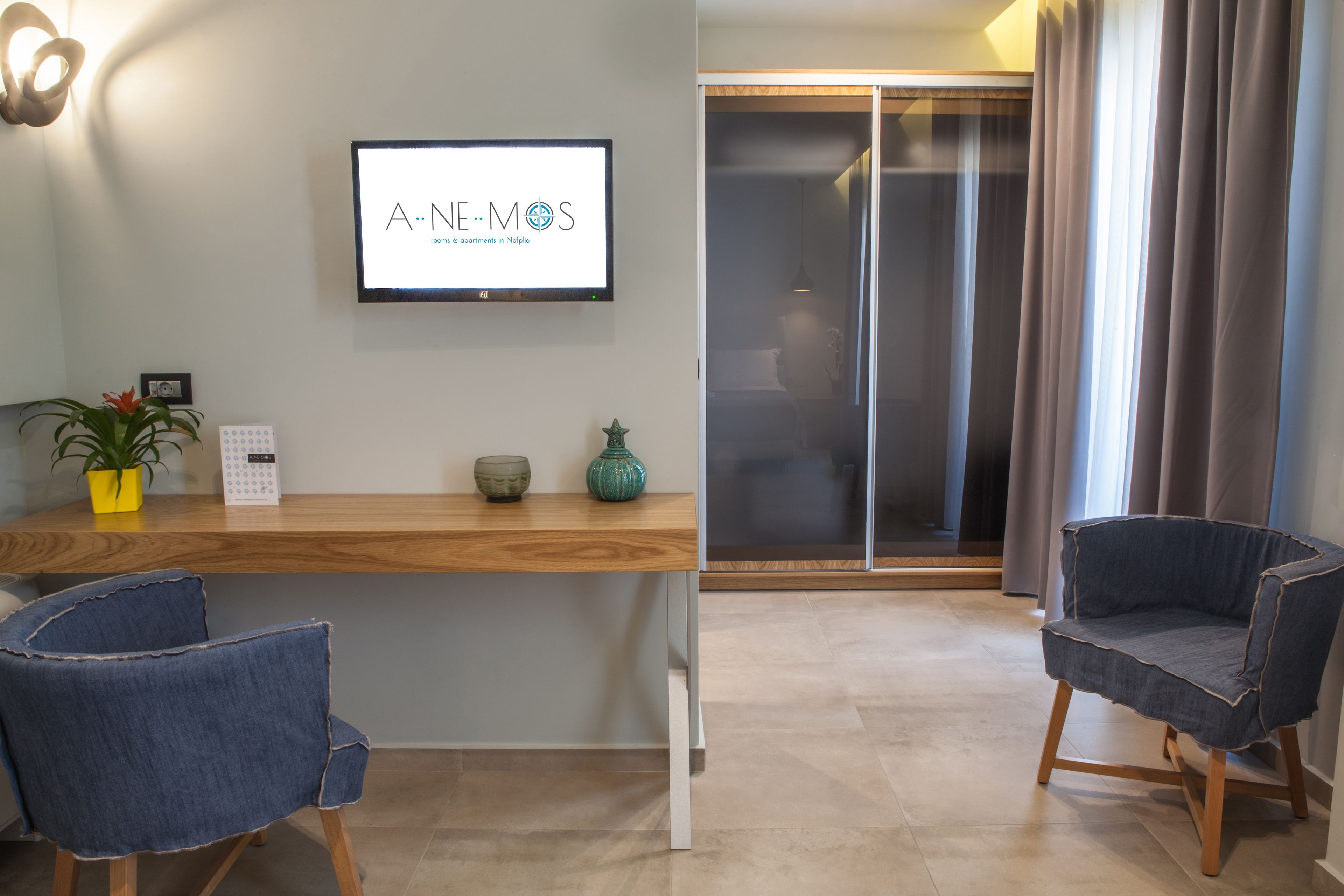 Habitació moderna amb escriptori de fusta, dues cadires de teixit blau, televisió que mostra “A.N.E.M.O.S”, una planta en test i una porta corredissa de vidre que dona a un balcó.