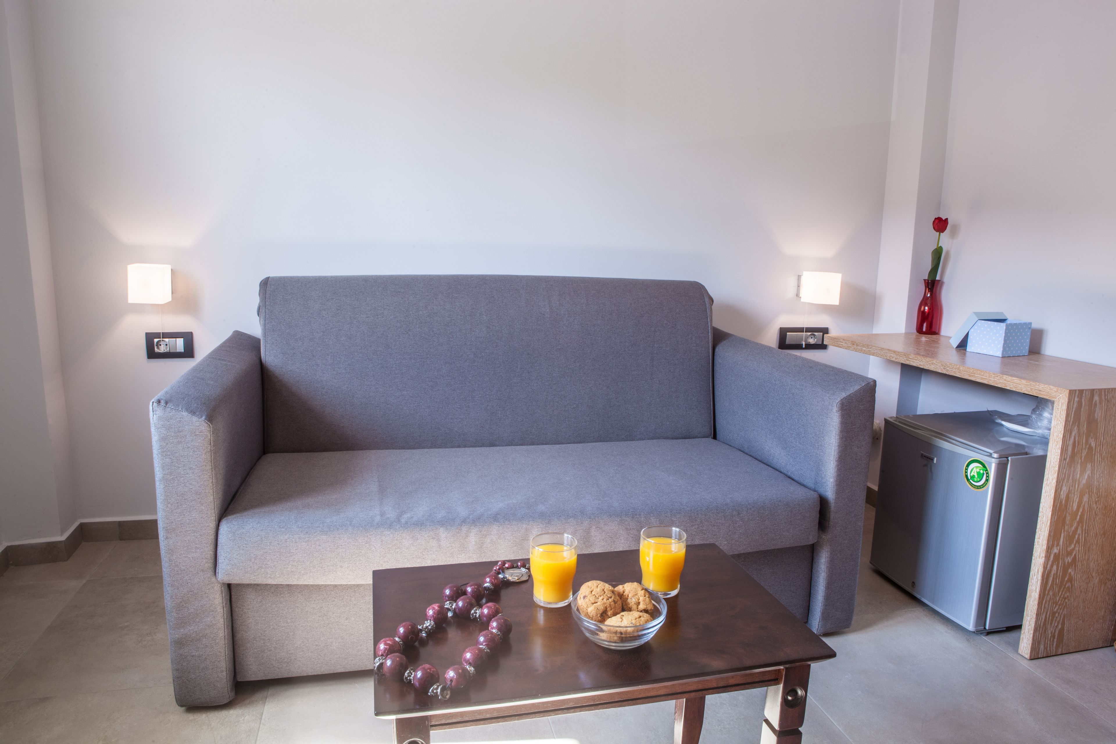 A grey sofa with two wall-mounted lamps, a small table with a bowl of cookies and glasses of orange juice, and a compact refrigerator.