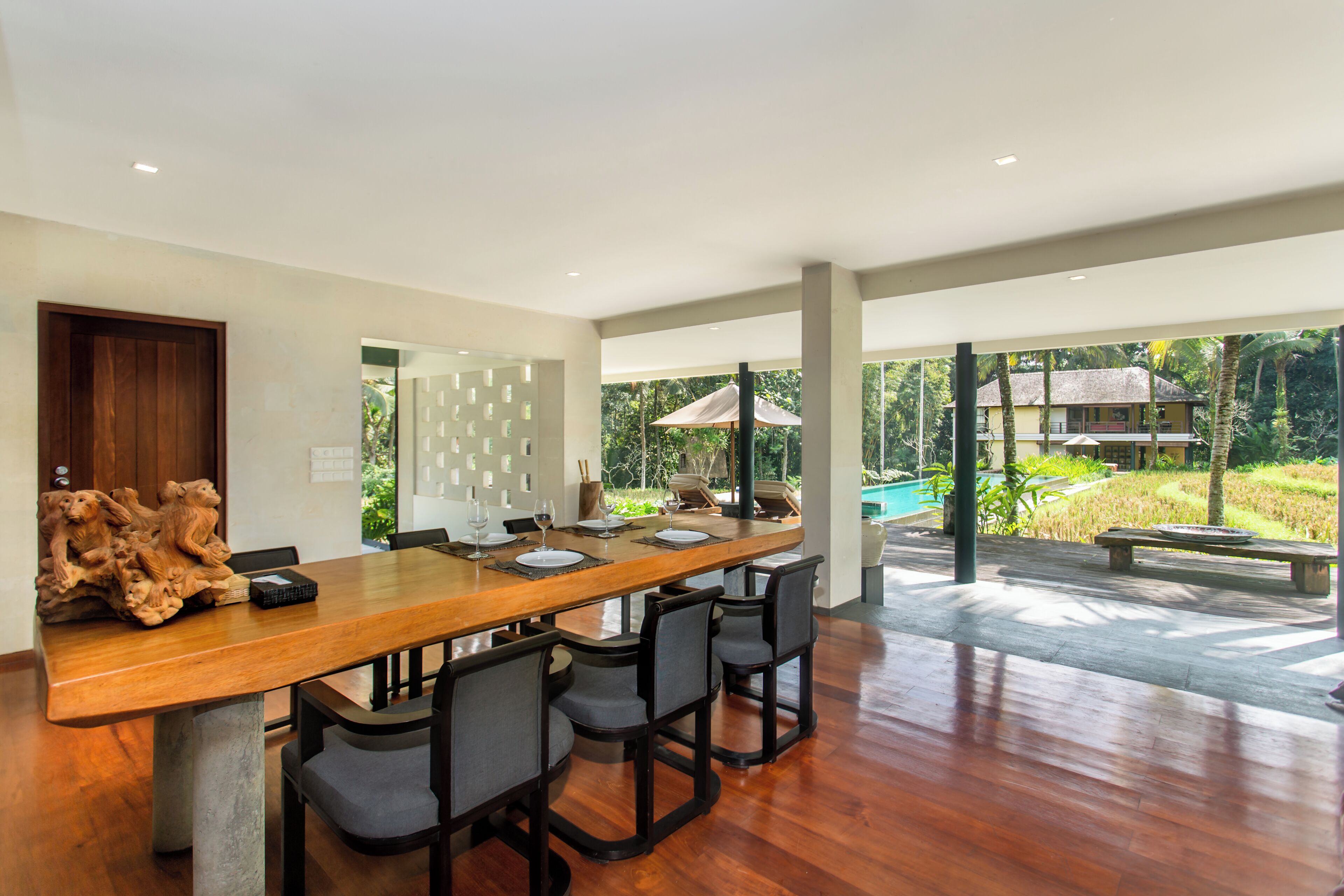 A spacious dining area with a long wooden table, multiple chairs, and a view of a swimming pool and lush greenery.