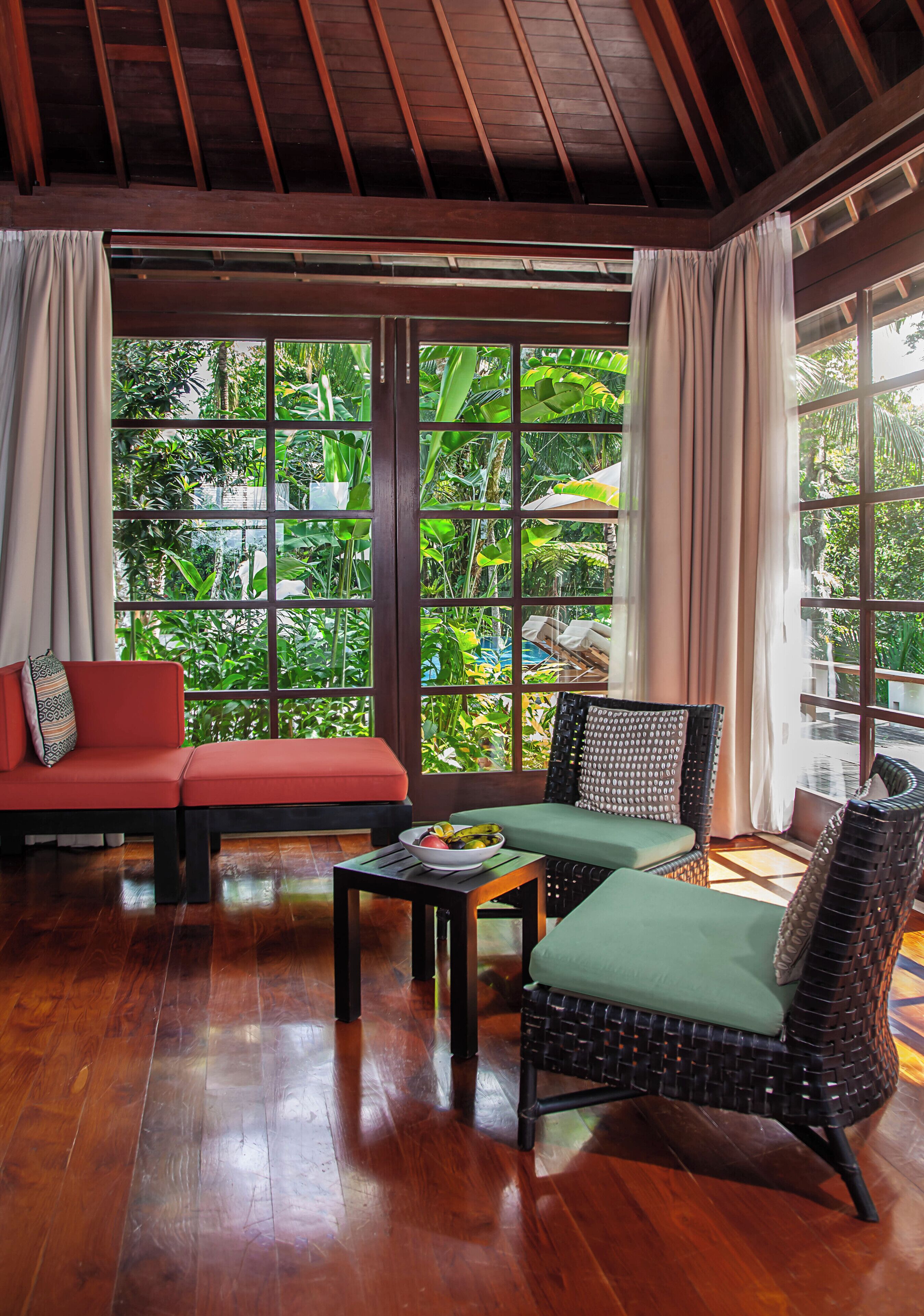 A room with a wooden ceiling, large windows with curtains, a red sofa, a wicker chair, and a small table with a bowl.