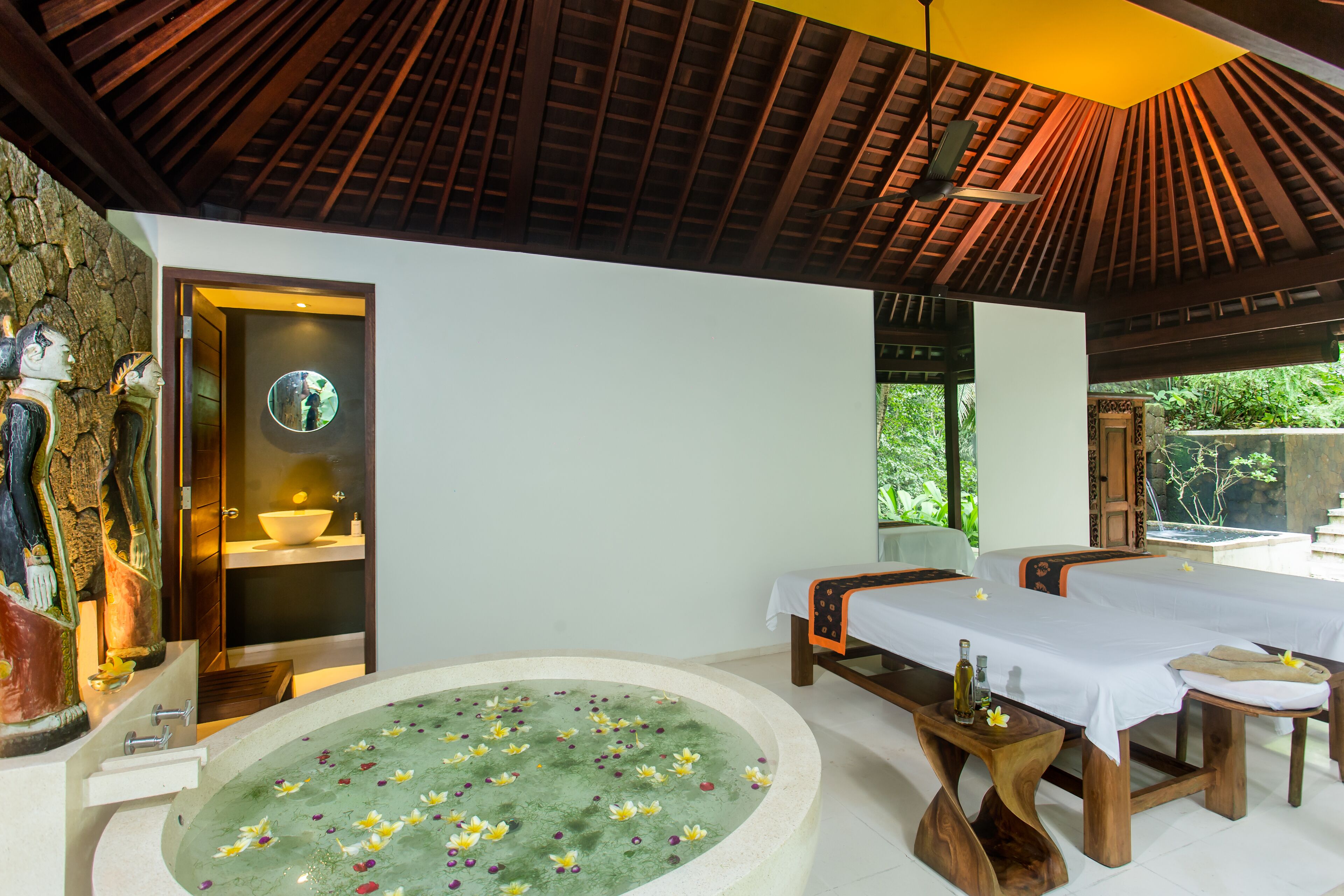 Une salle de spa dotée d’une grande baignoire remplie de fleurs, de deux tables de massage et d’un mur en pierre orné de sculptures.