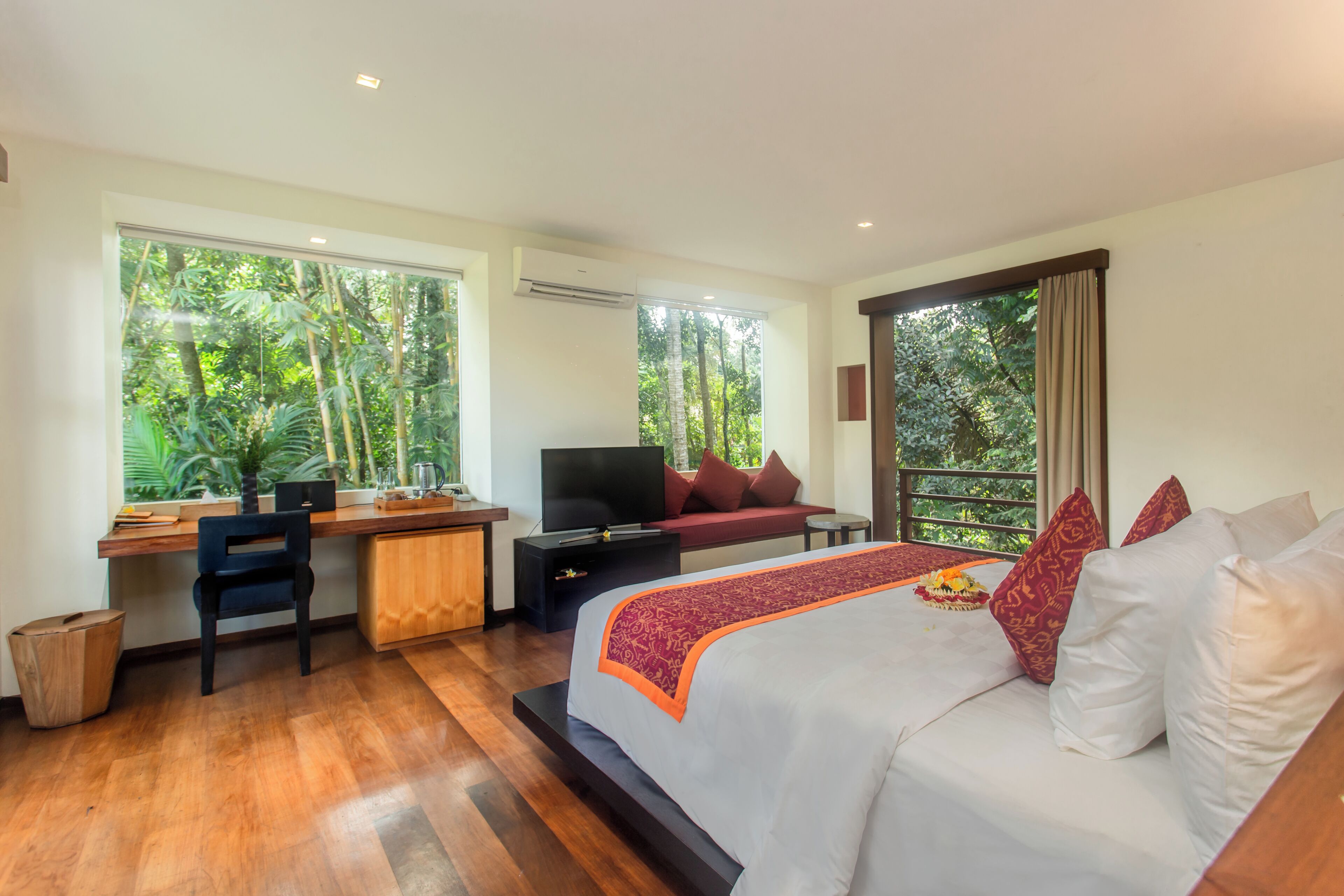 A bedroom with a bed, desk, and TV, large windows with a view of trees, and a balcony.