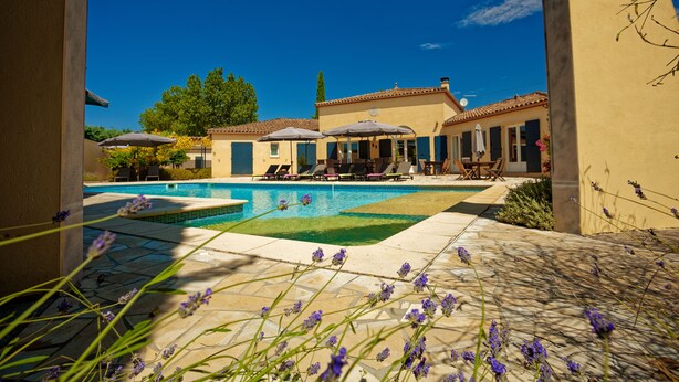 A pool area with lounge chairs and umbrellas, surrounded by a building with a tiled roof. - Villa des Cammaous (Vacquieres)
