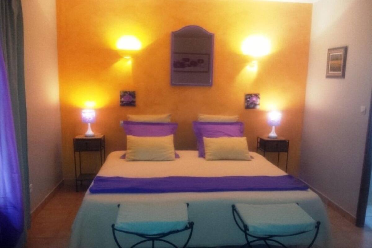 A bedroom with a bed, two bedside tables, two wall-mounted lamps, a framed picture, and two chairs.