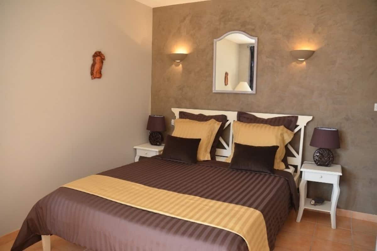 A neatly made bed with a brown and beige bedspread, two bedside lamps, a wall-mounted mirror, and a small decorative item on the wall.