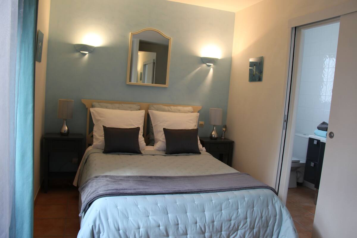 A neatly made bed with a headboard, two bedside tables with lamps, a wall-mounted mirror, and a door leading to a bathroom.