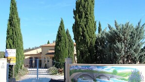 A gated entrance with a mural depicting a bridge and landscape, tall cypress trees, and a building with a tiled roof. - Villa des Cammaous (Vacquieres)