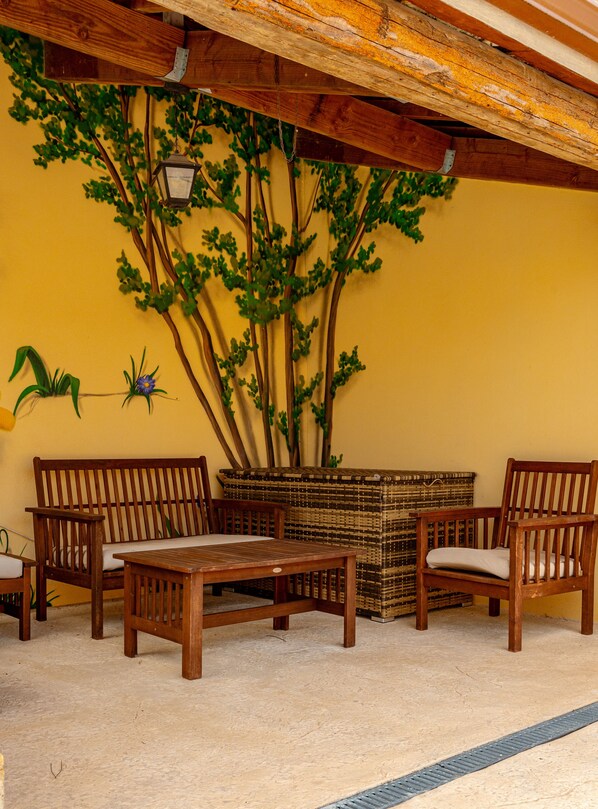 Un conjunto de banco y mesa de madera frente a una pared amarilla, con árboles artificiales y una cesta de mimbre.