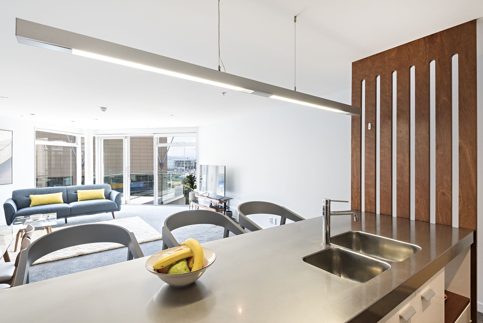 A modern kitchen with a stainless steel countertop, a bowl of fruit, and a view of the living area with a sofa and chairs.