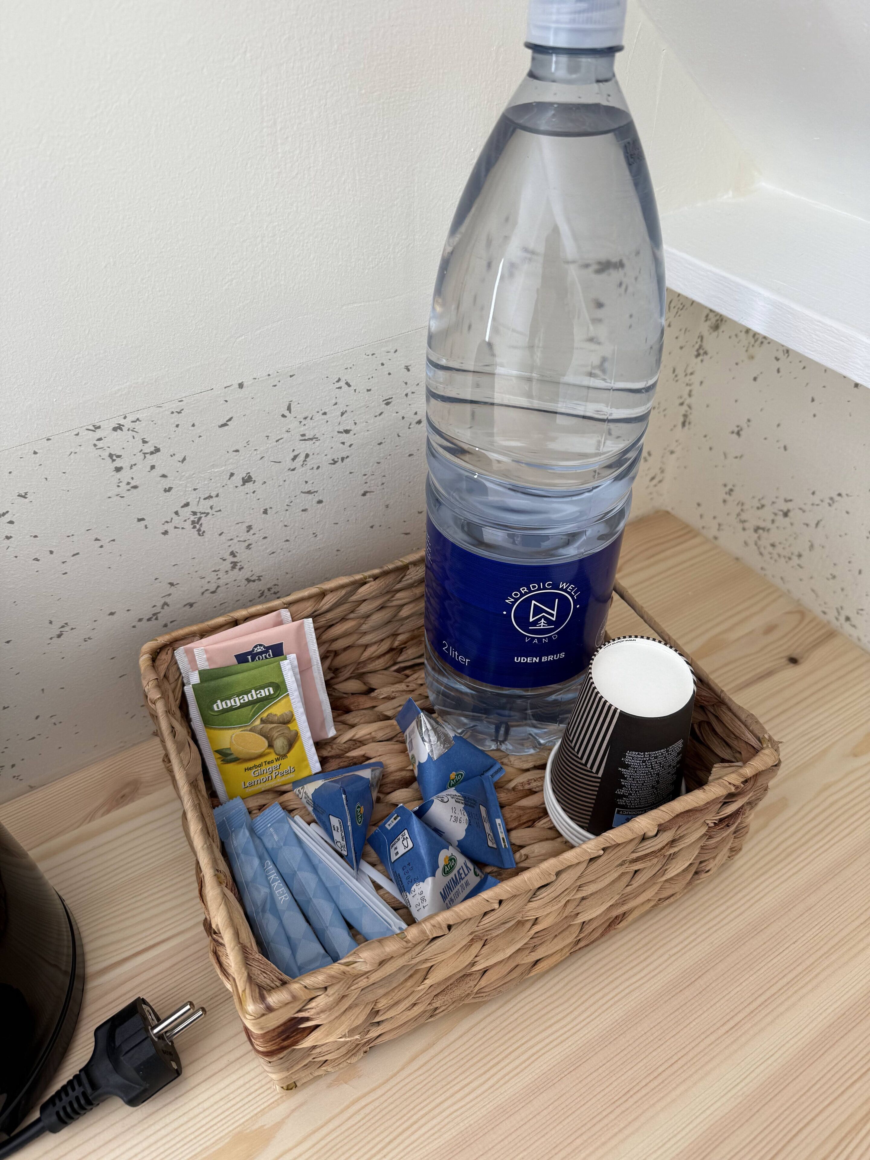 Ein Korbwägelchen mit einer Wasserflasche, Kräuterteesäckchen, einer Tasse und einem Netzteil auf einer Holztischplatte.