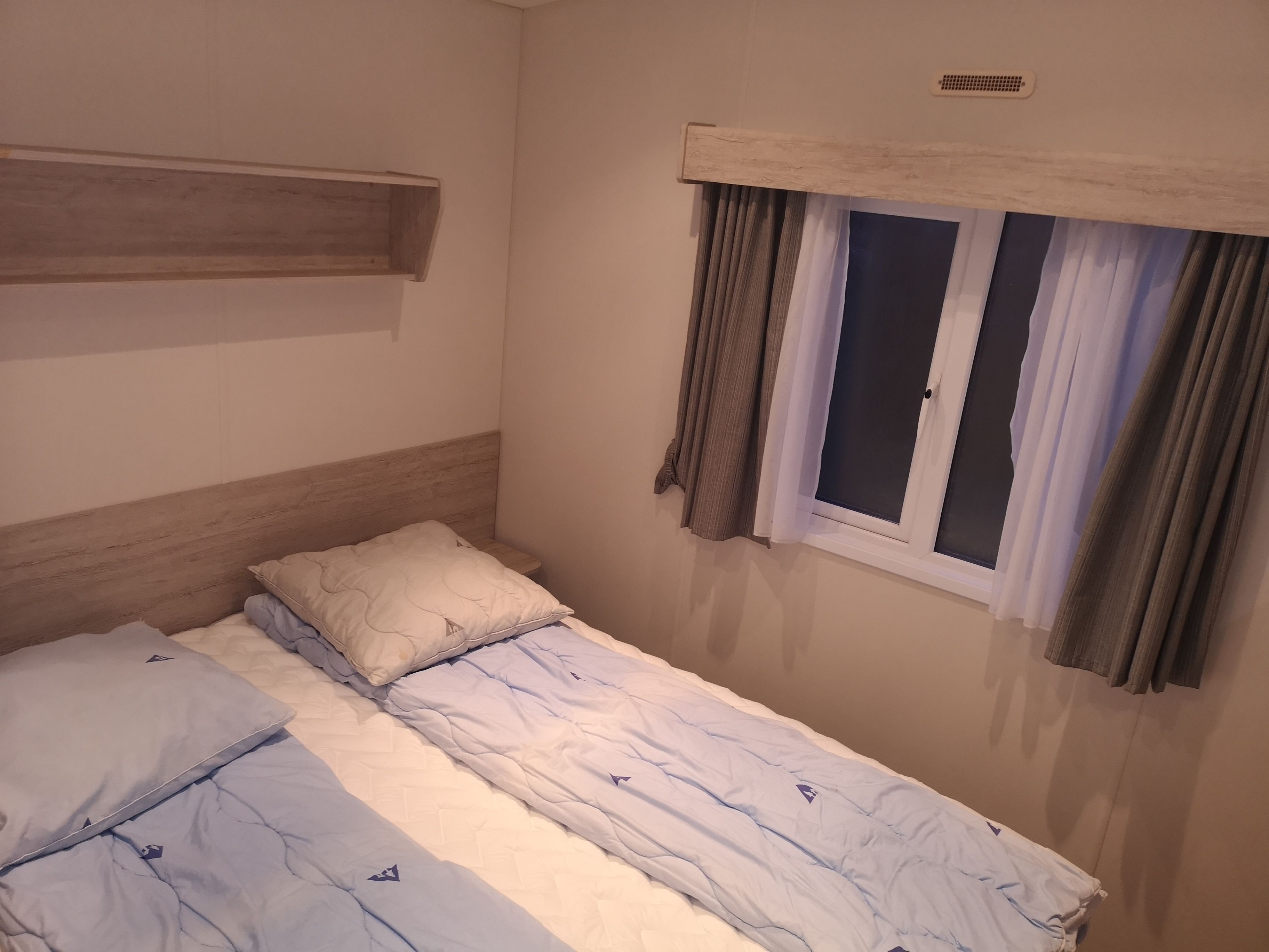 A single bed with a wooden headboard, a window with curtains, and a small shelf above the bed.