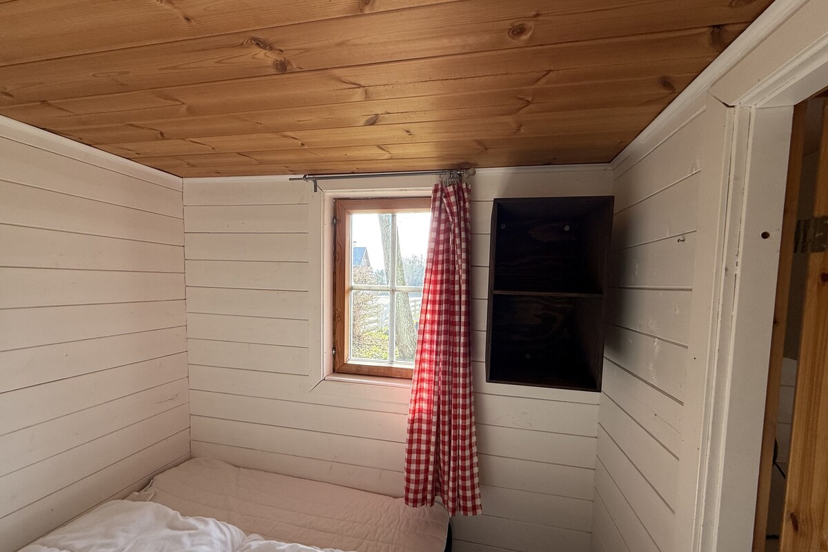 Une petite chambre avec un lit simple, des murs en bois et un plafond en pente. On y trouve une fenêtre avec un rideau à carreaux rouges et blancs, ainsi qu’une étagère le long du mur.