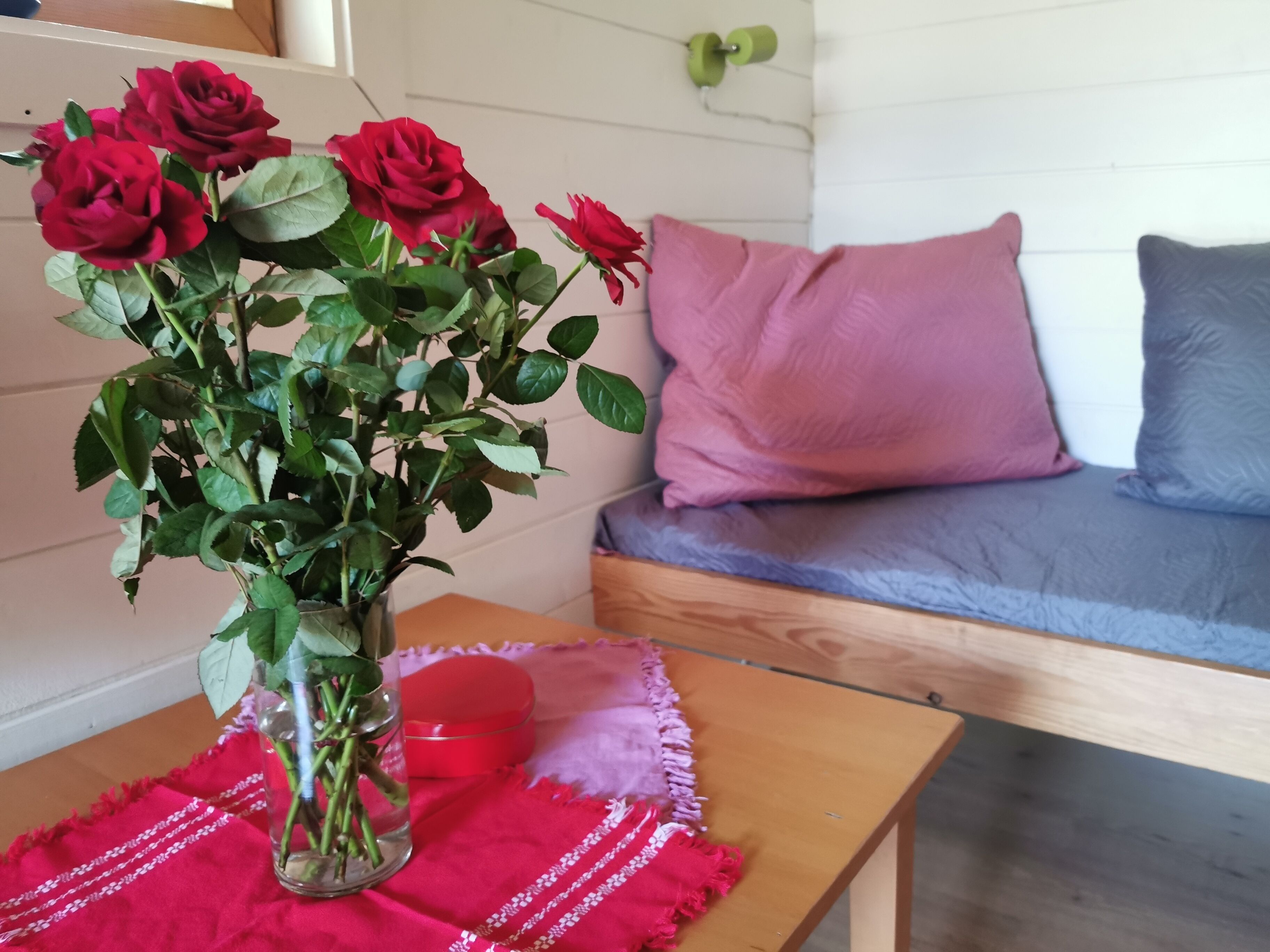 A room with a wooden table, a glass vase with red roses, a red cushion, and a grey sofa with pink and grey cushions.