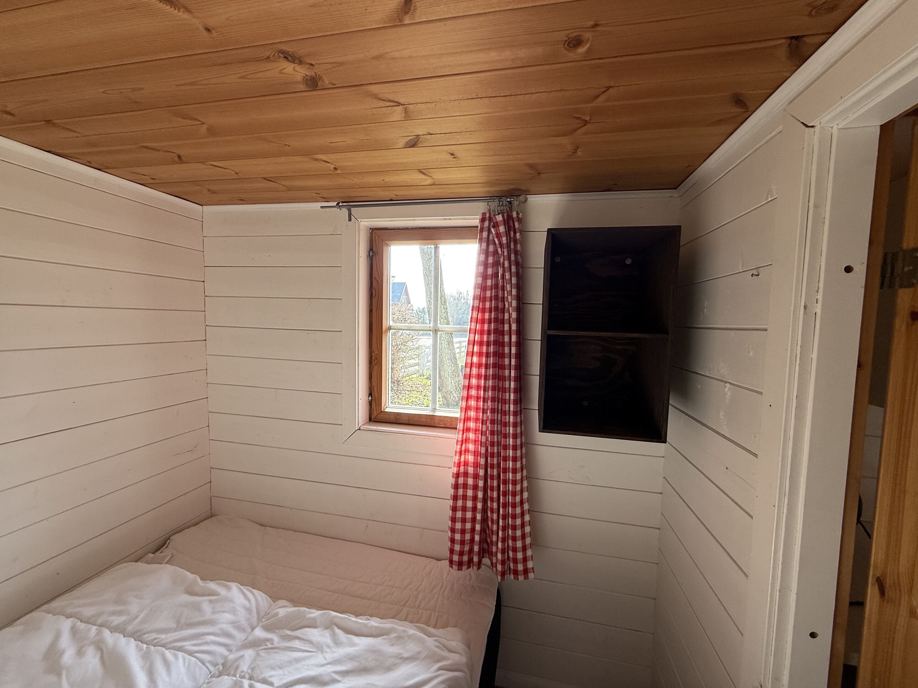 A small, single-bed room with wooden walls and a sloped ceiling. There is a window with a red and white checkered curtain, and a shelf unit against the wall.