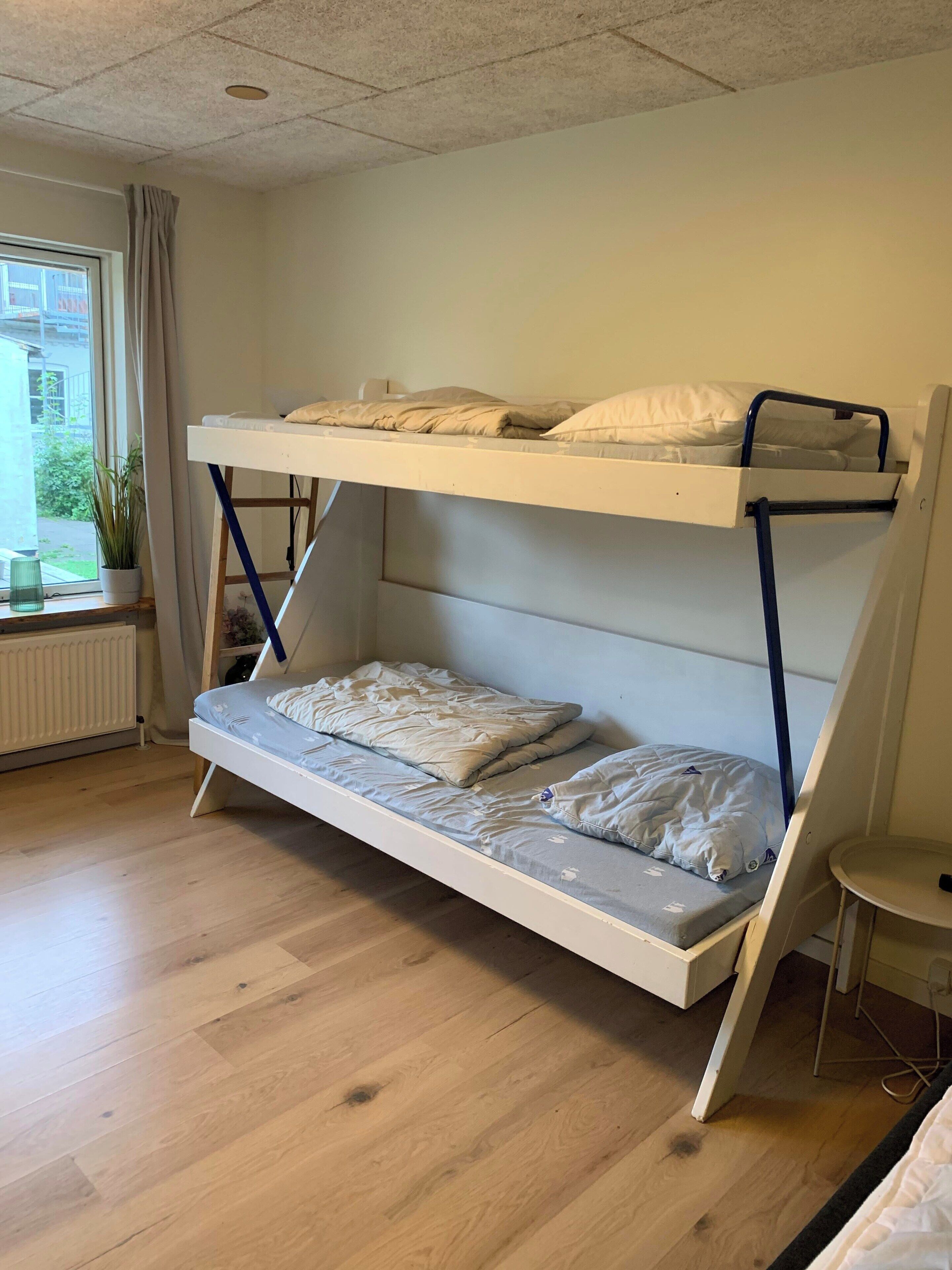 Egy emeletes ágy, fehér kerettel, alsó ágyrésze matraccal és párnákkal, valamint egy ablak, amelyen függöny van.
