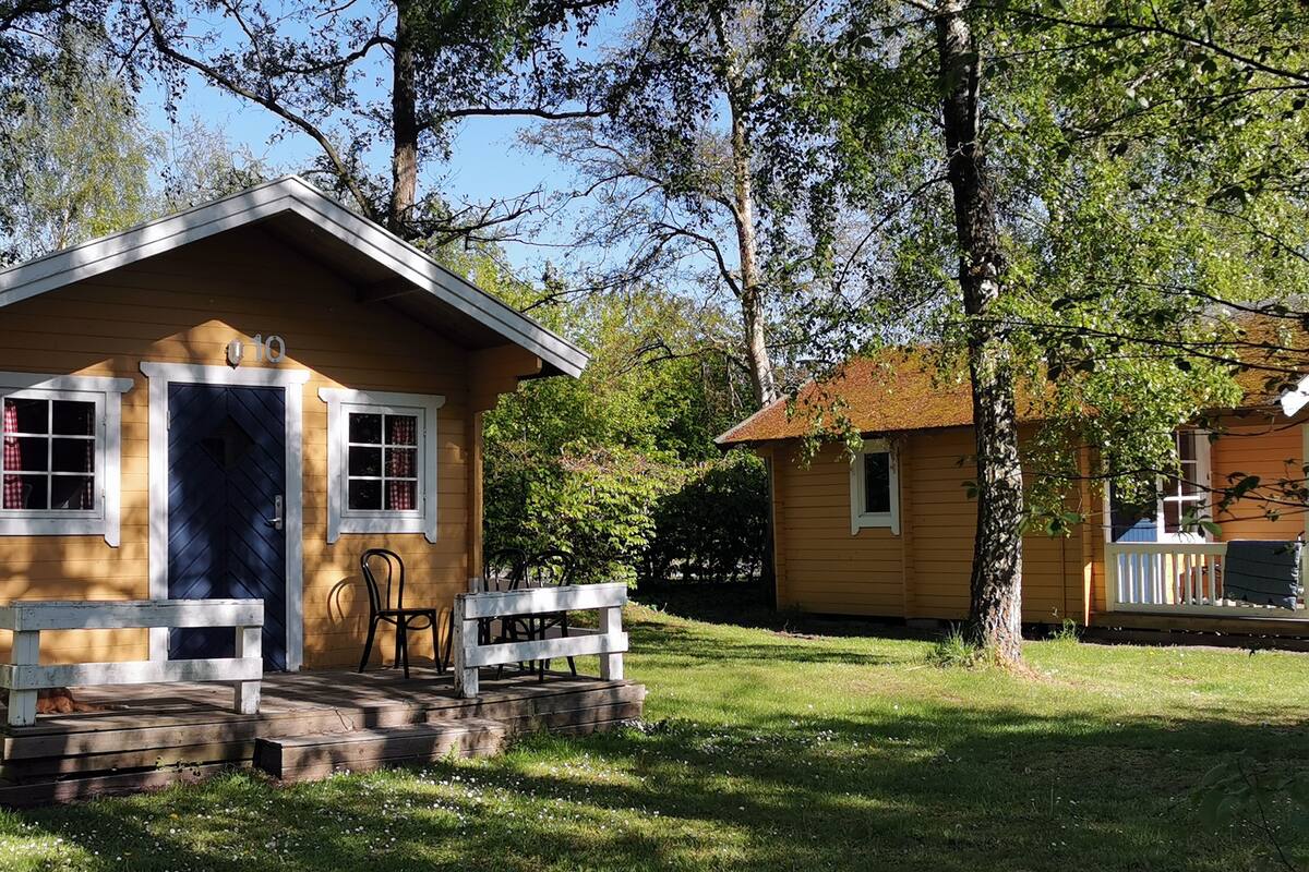 Una cabaña de madera pequeña con un porche y una pequeña terraza, rodeada de árboles y césped.