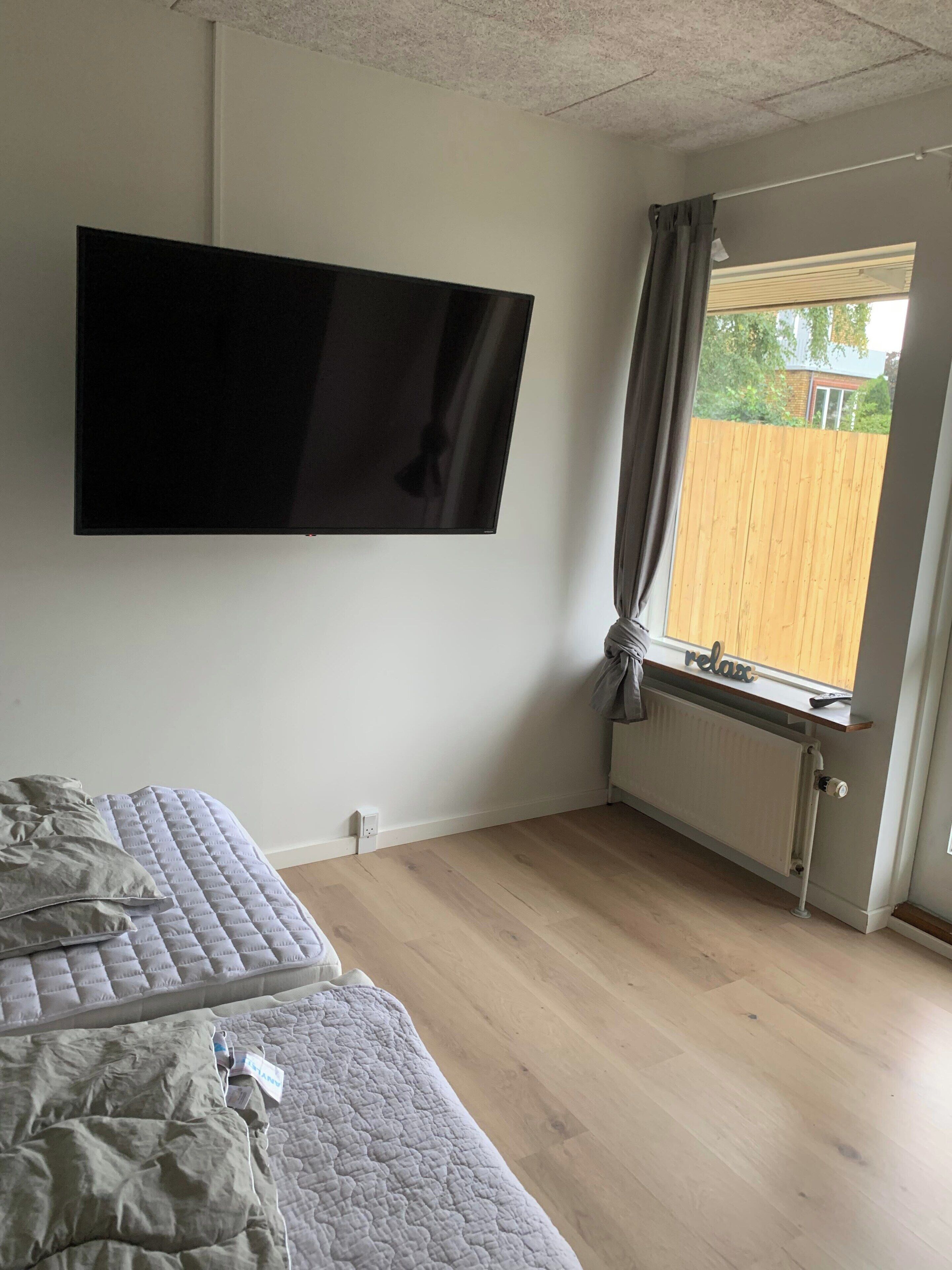Une chambre à coucher avec un lit, un téléviseur fixé au mur et une fenêtre avec des rideaux.