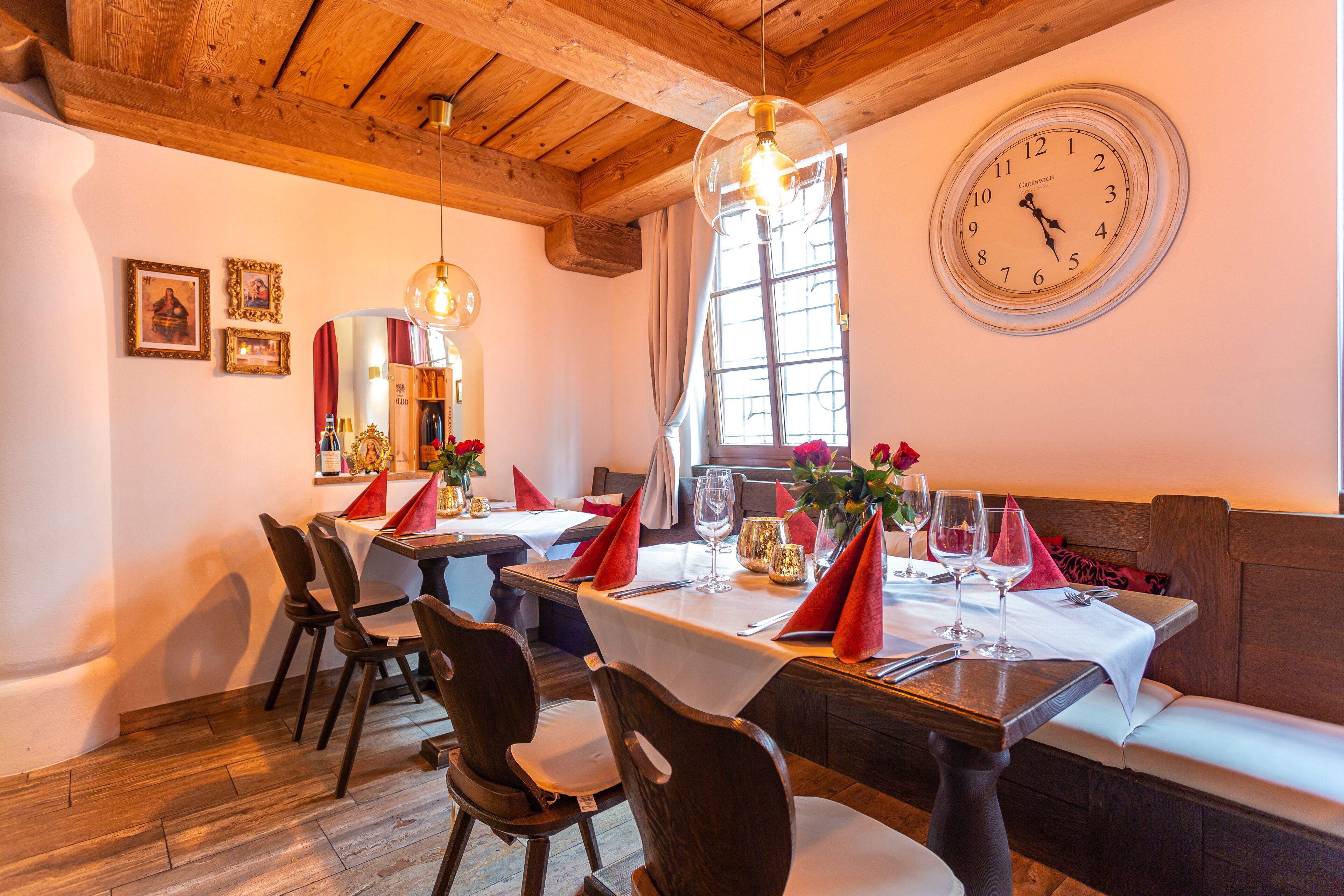 A restaurant with wooden beams, a large wall clock, and tables set for dining with red napkins.