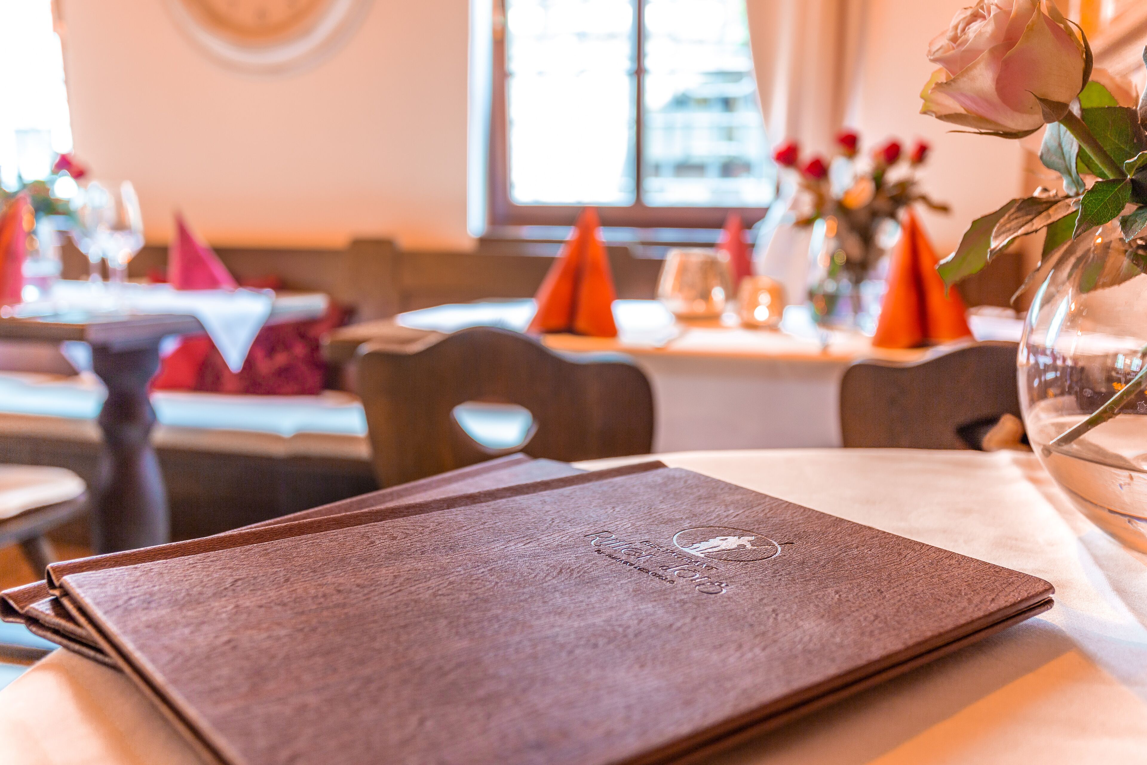 A restaurant table with a menu, a vase with a rose, and a window in the background.