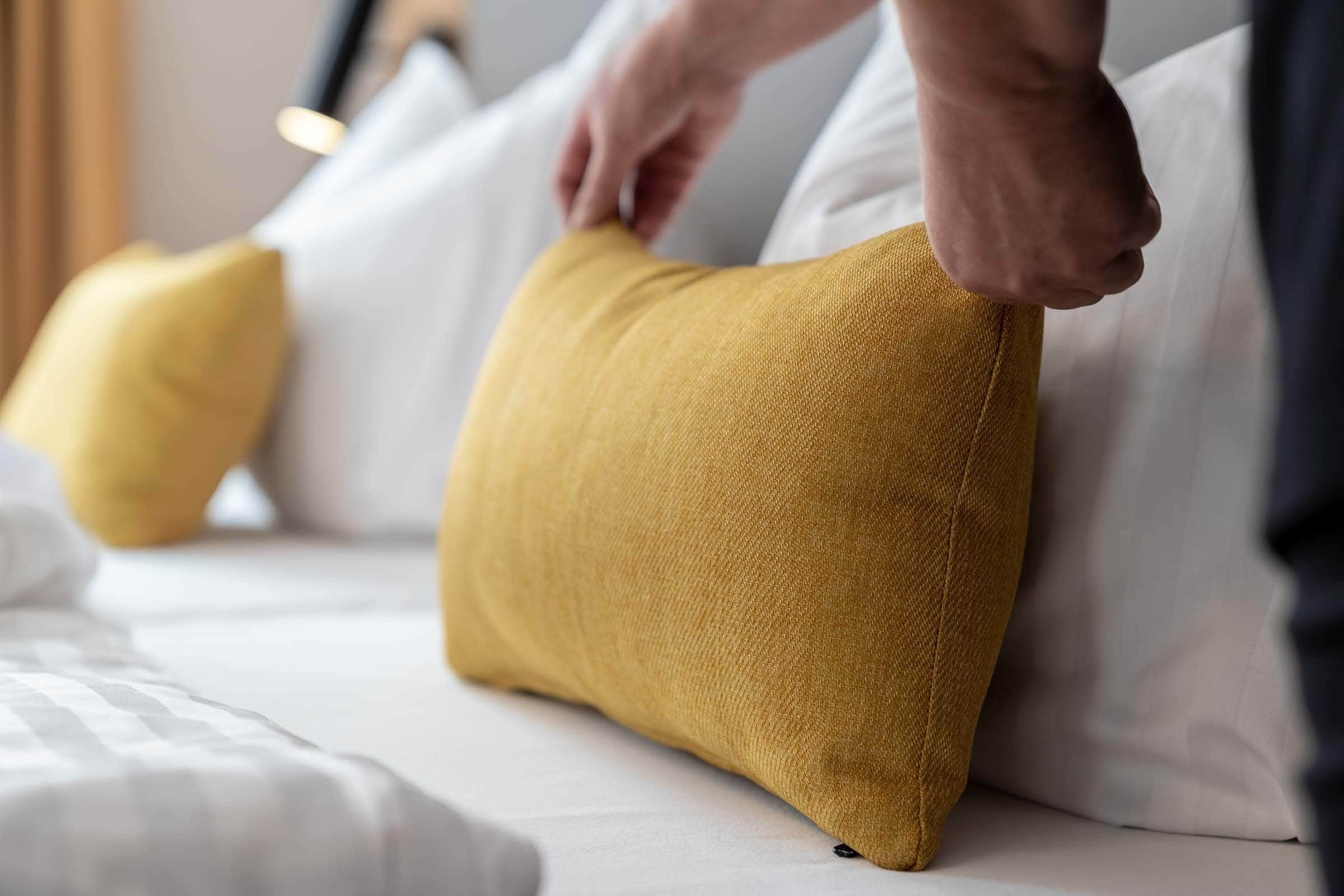 Une personne ajuste un coussin de couleur moutarde sur un lit recouvert de draps blancs.