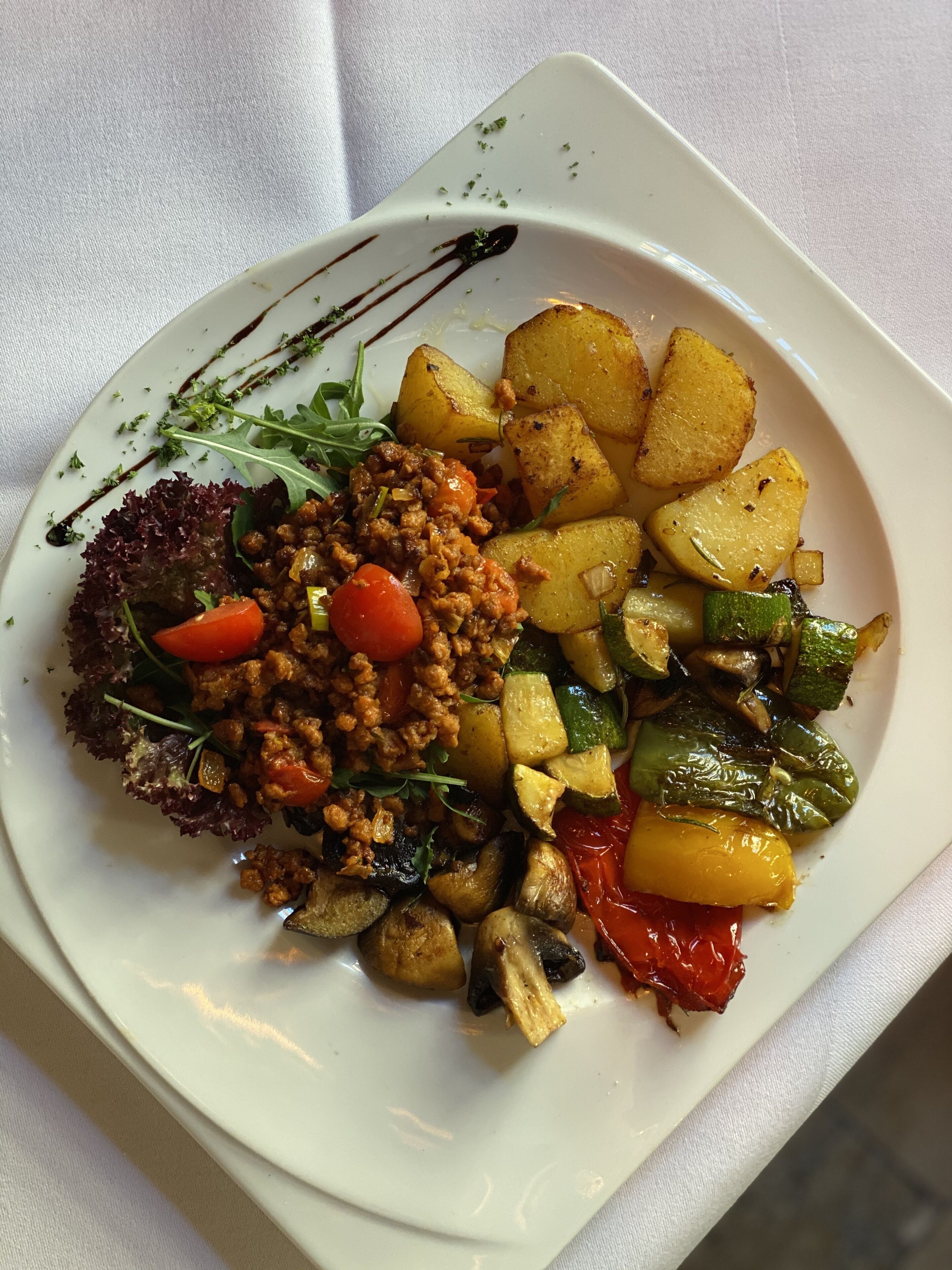 Un plato cuadrado blanco con diversos alimentos, incluyendo una hamburguesa, papas asadas y verduras mixtas.