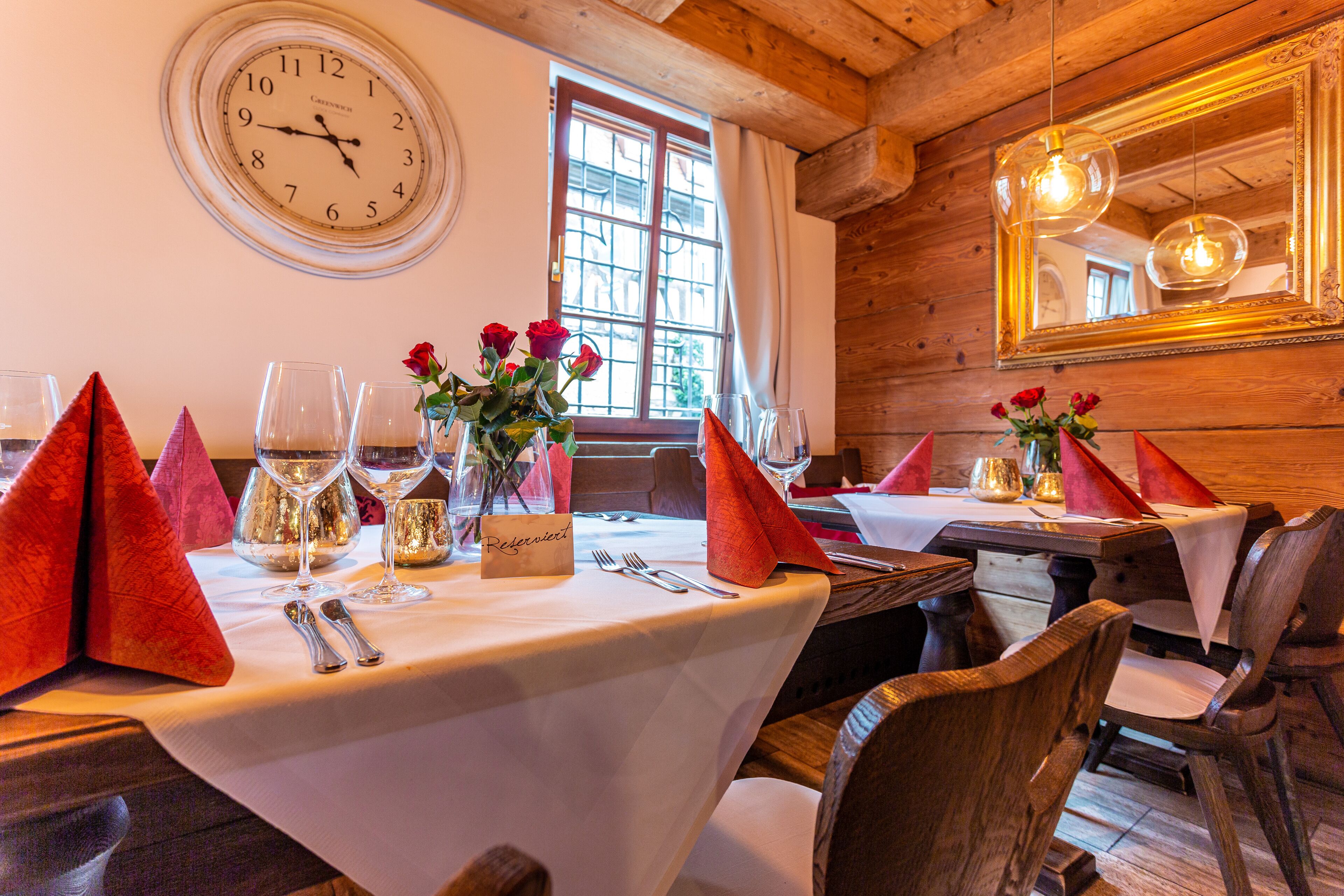 A dining table set with red napkins, wine glasses, and a vase of red roses. A clock and a mirror are visible on the wall.