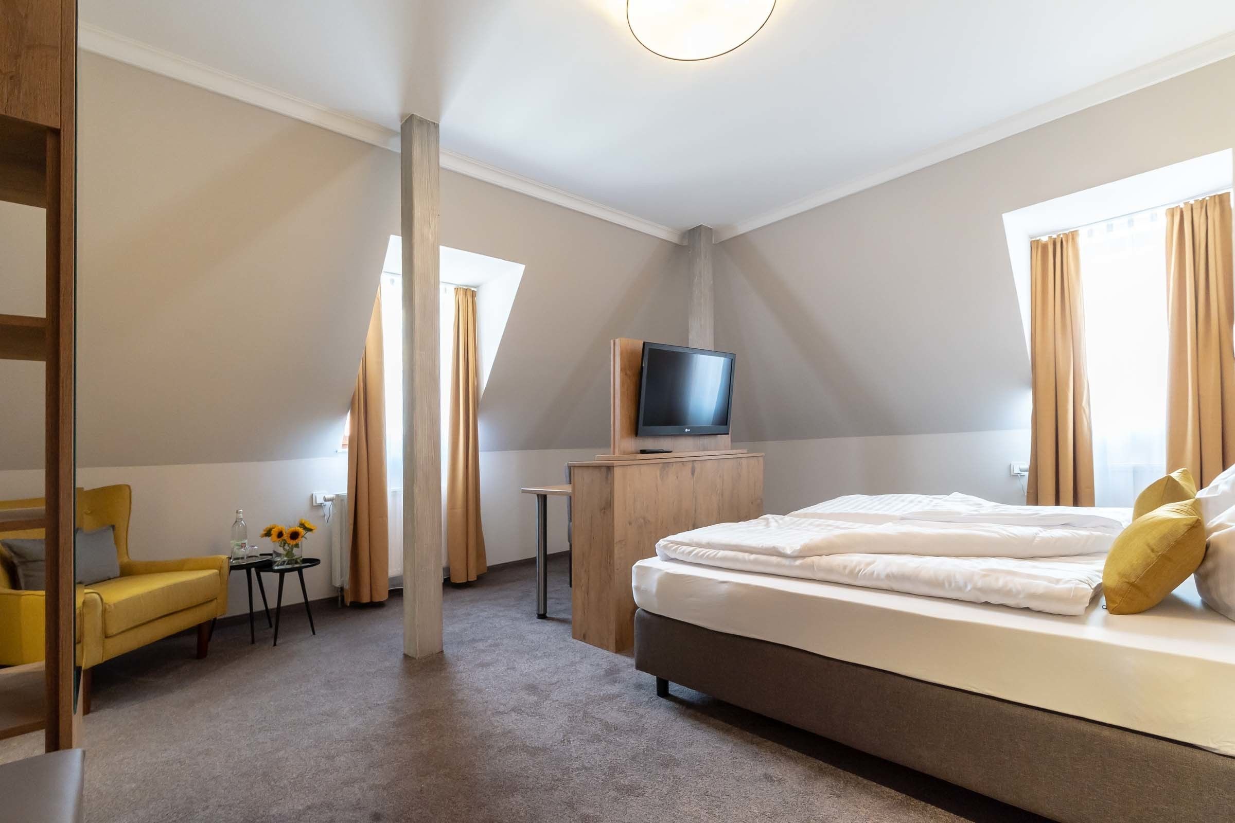 Habitación de hotel con una cama, un televisor sobre un mueble de madera, un sillón amarillo, una pequeña mesa con un jarrón de girasoles y una ventana con cortinas.