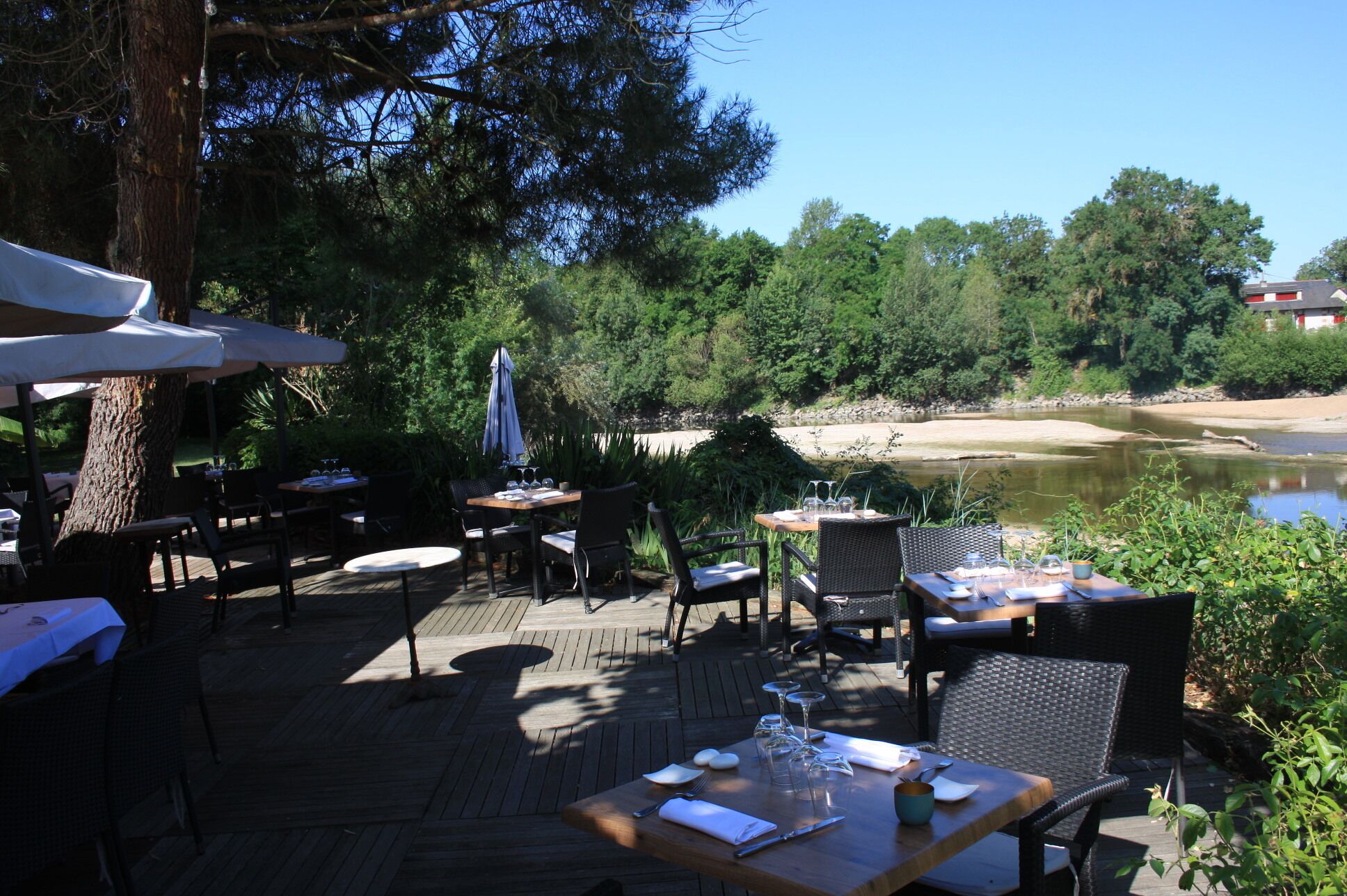 An outdoor dining area with tables and chairs, surrounded by trees and a view of a river.