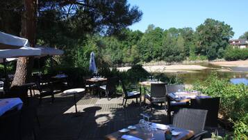 An outdoor dining area with tables and chairs, surrounded by trees and a view of a river.