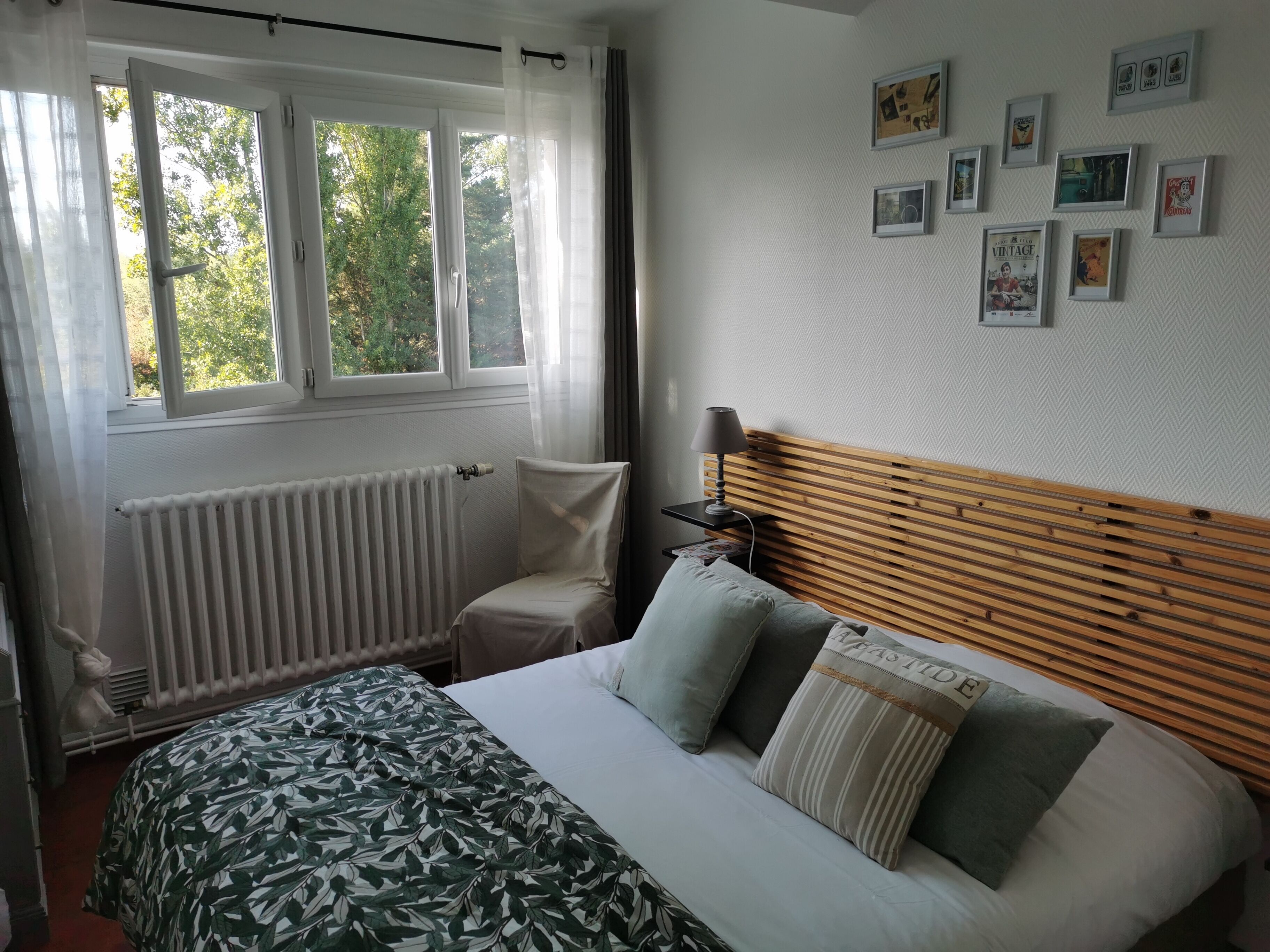 A bedroom with a bed, a radiator, a window with curtains, and a wall with framed pictures.
