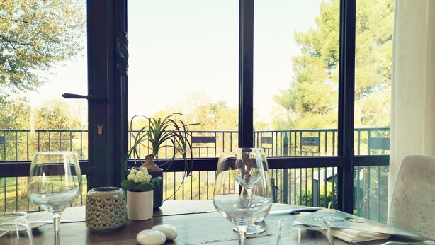 Une table à manger avec des verres à vin, un vase rempli de fleurs et une vue sur un balcon doté d’une balustrade et bordé d’arbres.