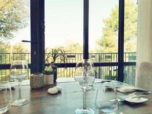 Een eettafel met wijnglazen, een vaas met bloemen en uitzicht op een balkon met een hekwerk en bomen.