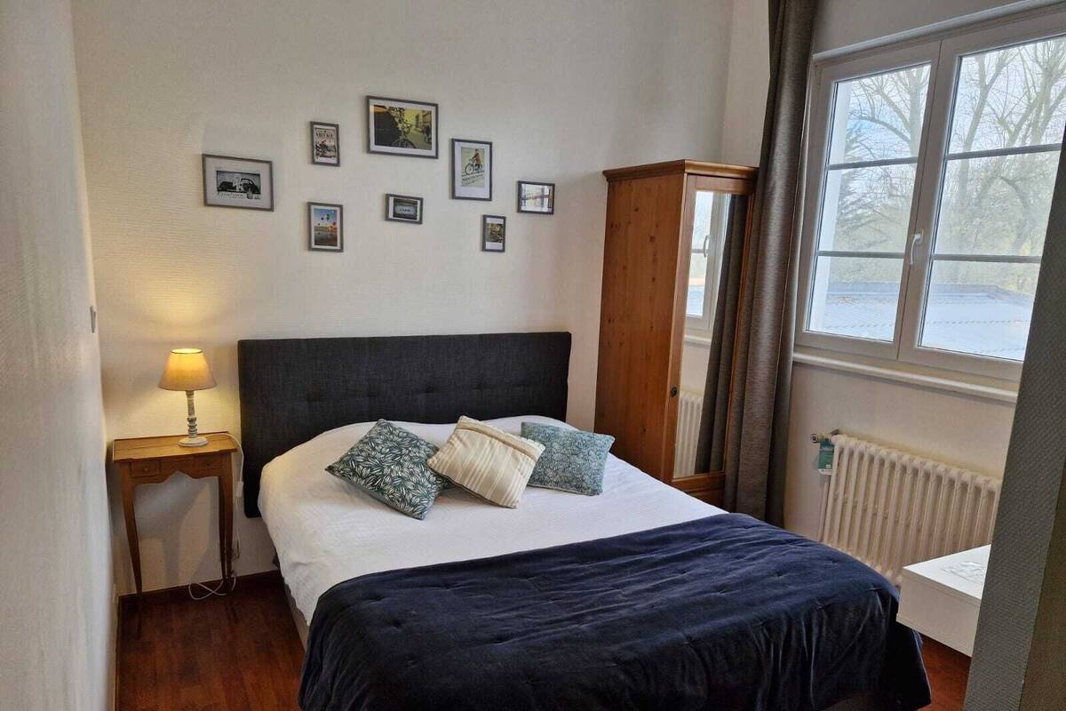 Un dormitorio con cama, mesita de noche, armario de madera, radiador y ventana con vistas al exterior.
