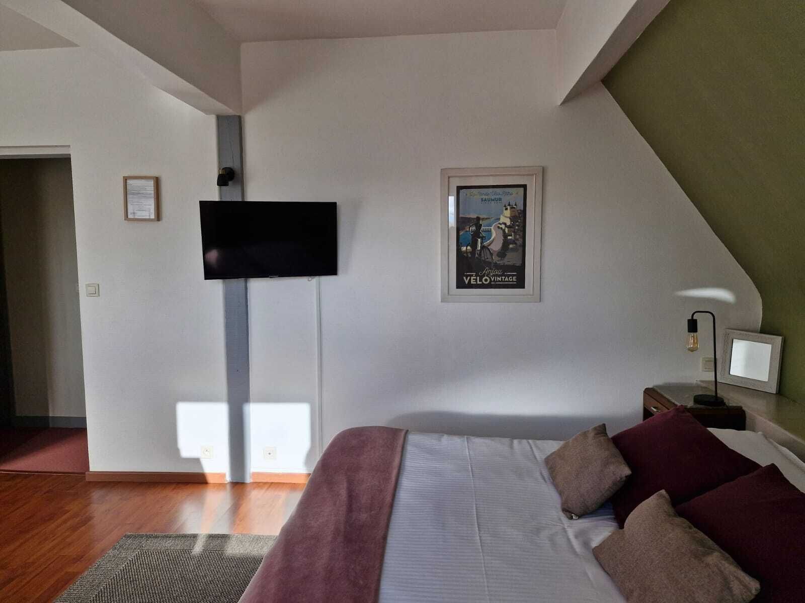 Un dormitorio con cama, almohadas, televisor montado, un cartel enmarcado y una lámpara de noche.