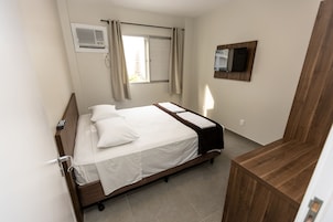Una habitación de hotel ordenada con una cama, una mesita de noche de madera, un televisor montado y una ventana con cortinas.
