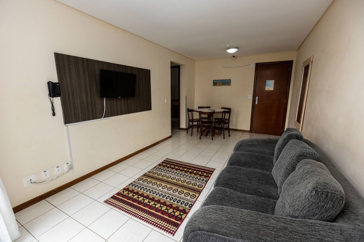 A living room with a grey sofa, a patterned rug, a flat-screen TV mounted on the wall, a dining table with chairs, and a door with a poster.