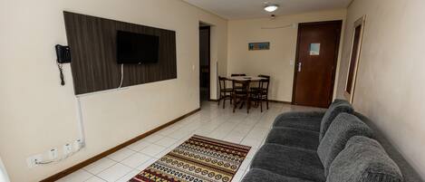 Una sala de estar con un sofá gris, un tapete estampado, un televisor de pantalla plana montado en la pared, una mesa de comedor con sillas y una puerta con un cartel.