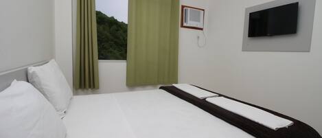 A hotel room with a bed, a window with curtains, an air conditioning unit, and a television mounted on the wall.