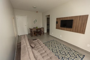 A living room with a sofa, a patterned rug, a TV mounted on a wooden panel, and a dining area with a table and chairs. - Hotel do Bosque (Balneário Camboriú)