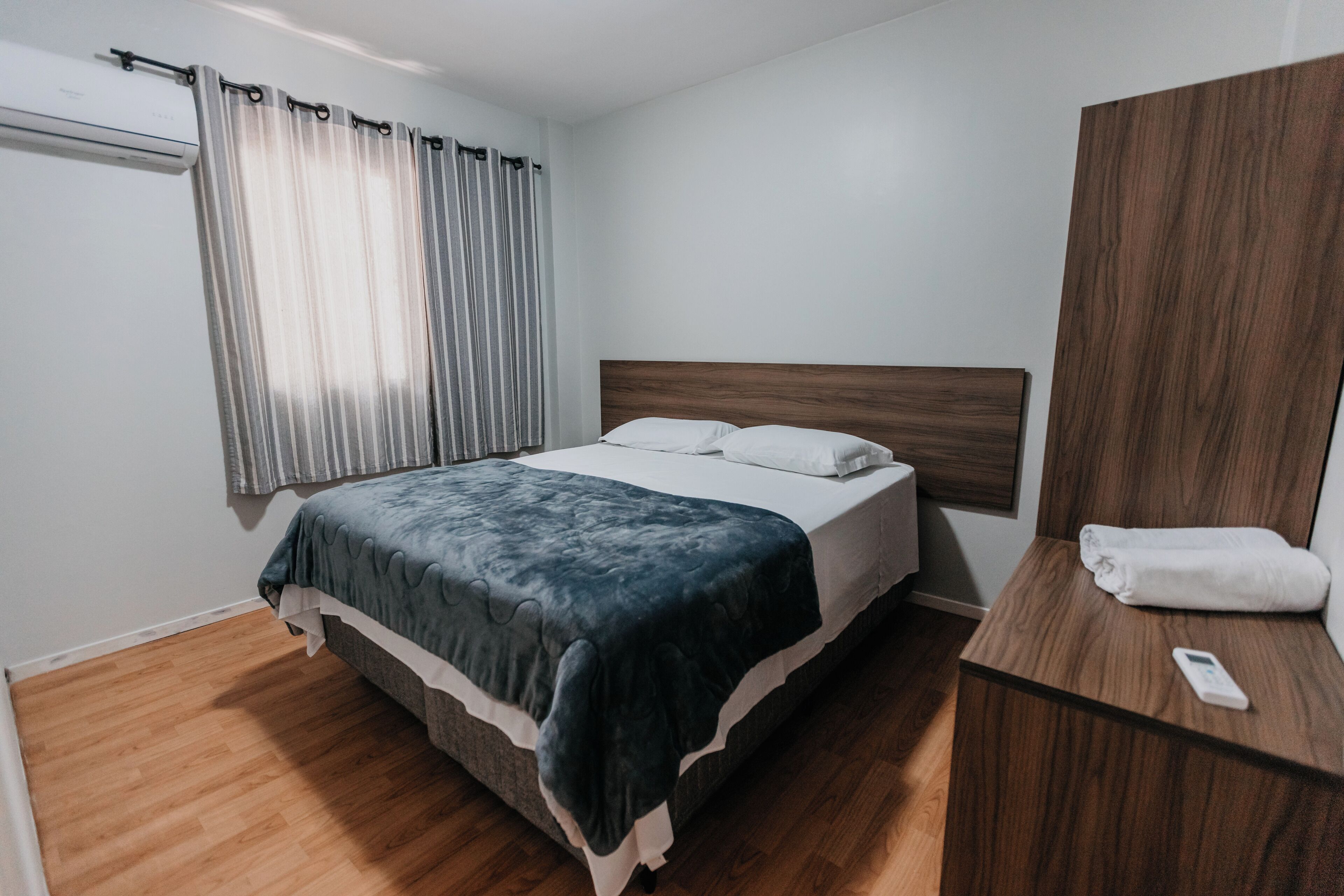 Quarto com cama, cabeceira de madeira, mesa de cabeceira com toalha e janela com cortinas.