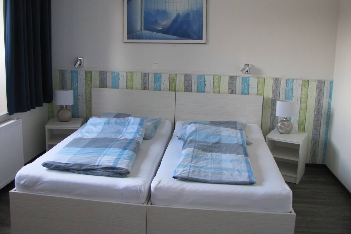 A double bed with a checkered bedspread, two bedside tables with lamps, a painting on the wall, and a window with curtains.