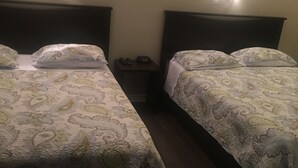 A hotel room with two beds, each with a patterned bedspread and pillows, a small bedside table with a phone and a lamp, and a framed picture on the wall. - Goodman's Motel (Baldwin)