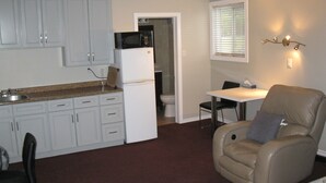 A compact kitchen with white cabinets, a refrigerator, and a microwave. There's a dining area with a table and chairs, and a leather armchair. - Goodman's Motel (Baldwin)